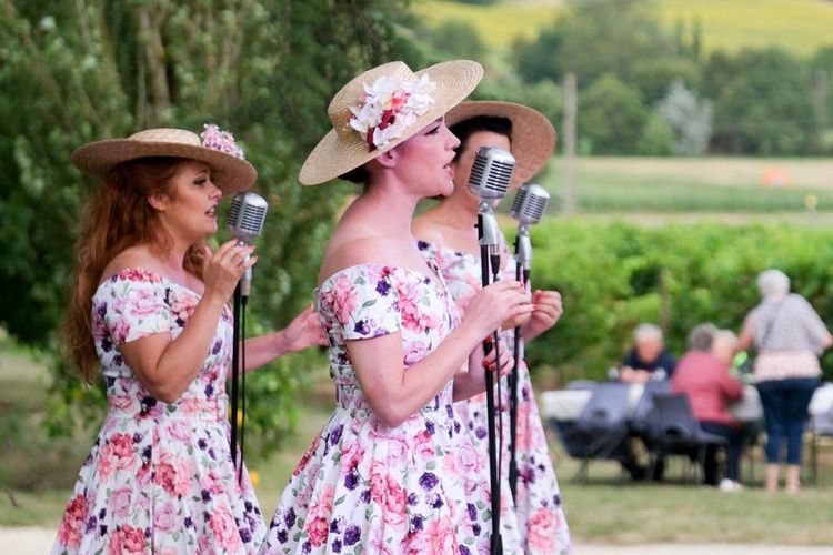 Trois femmes en train de chanter avec robes et chapeaux fleuris et des personnes en train de manger en arrière-plan avec des vignes